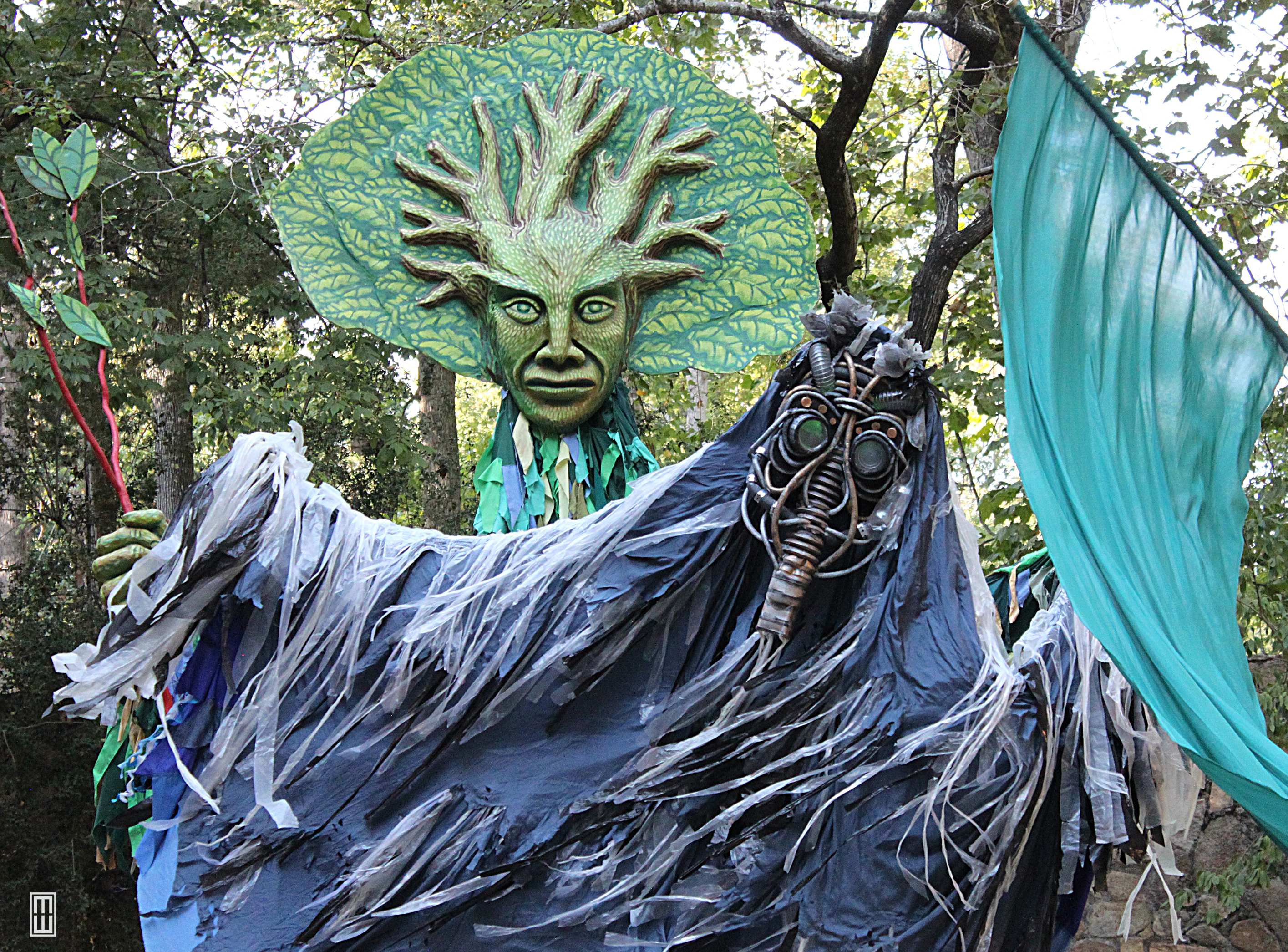 A "pollution monster" puppet with a ragged gray body and a gas mask-like face drifts in front of a tree-headed forest god puppet.