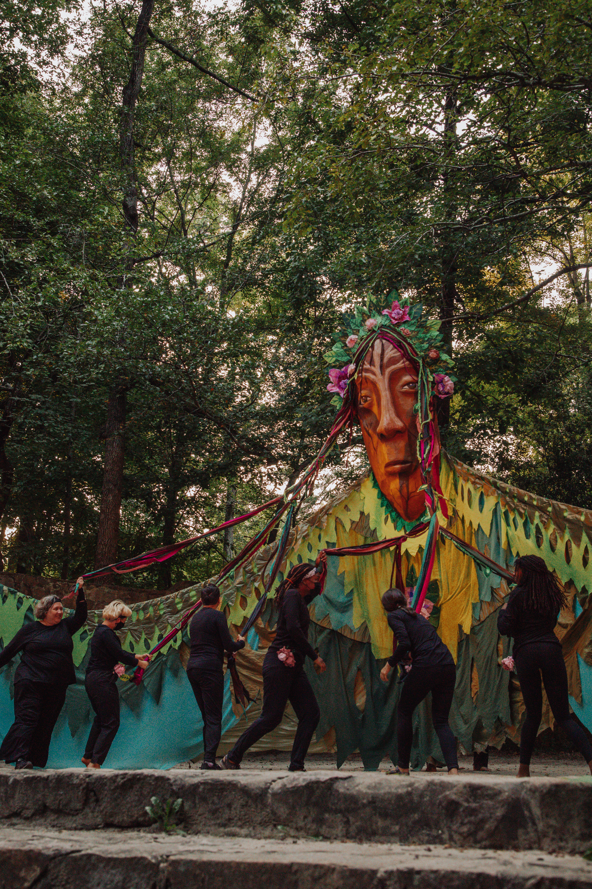 A 20 foot tall Earth goddess puppet, wearing draping green clothing and crowned with flowers. 6 performers are in the midst of a dance where they braid her multcolored hair.