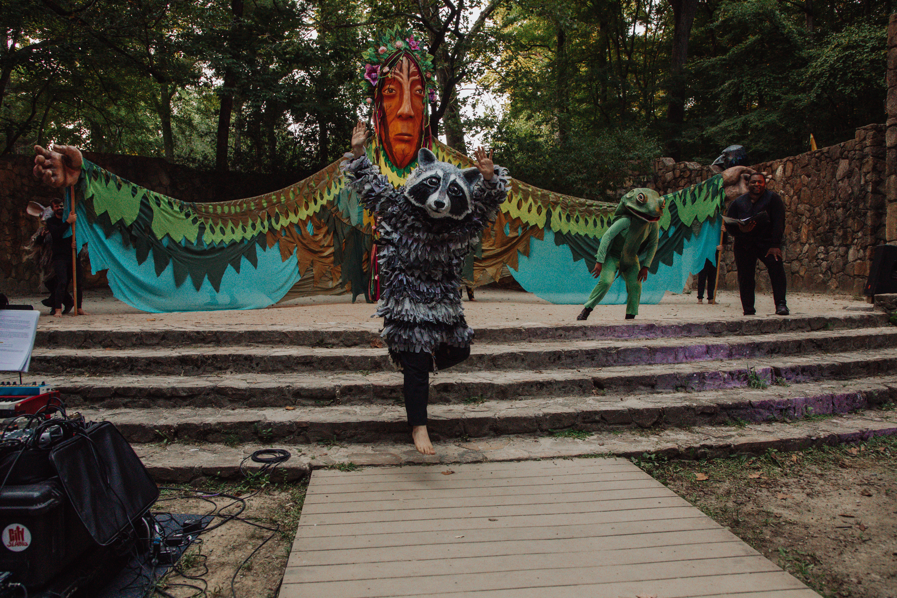 Xtian dressed in a raccoon costume leaps off a stage. A 20 foot tall goddess puppet stands in the background.