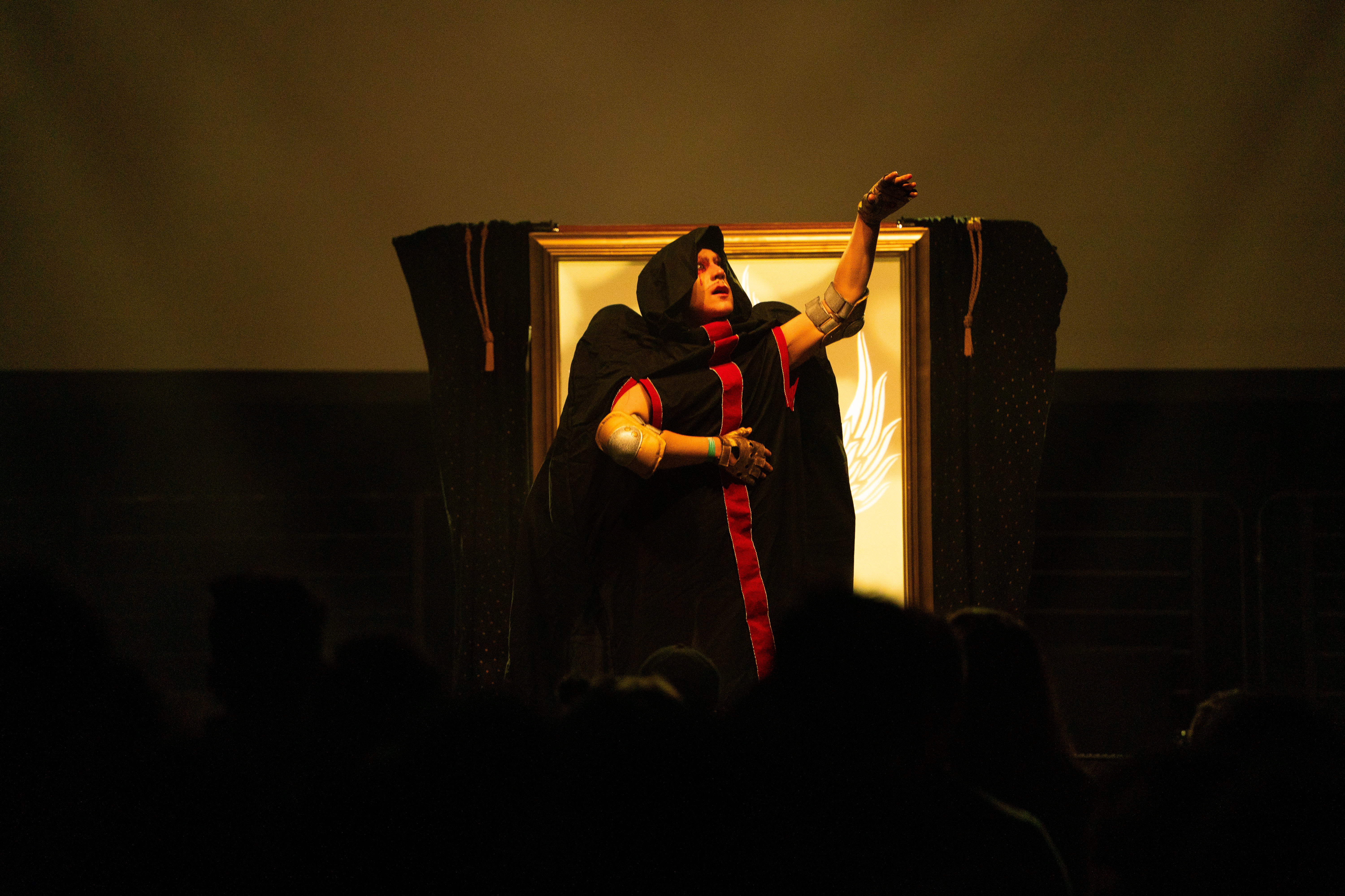 Xtian Mingle stands on stage in a red and black cloak, reaching up with one hand.