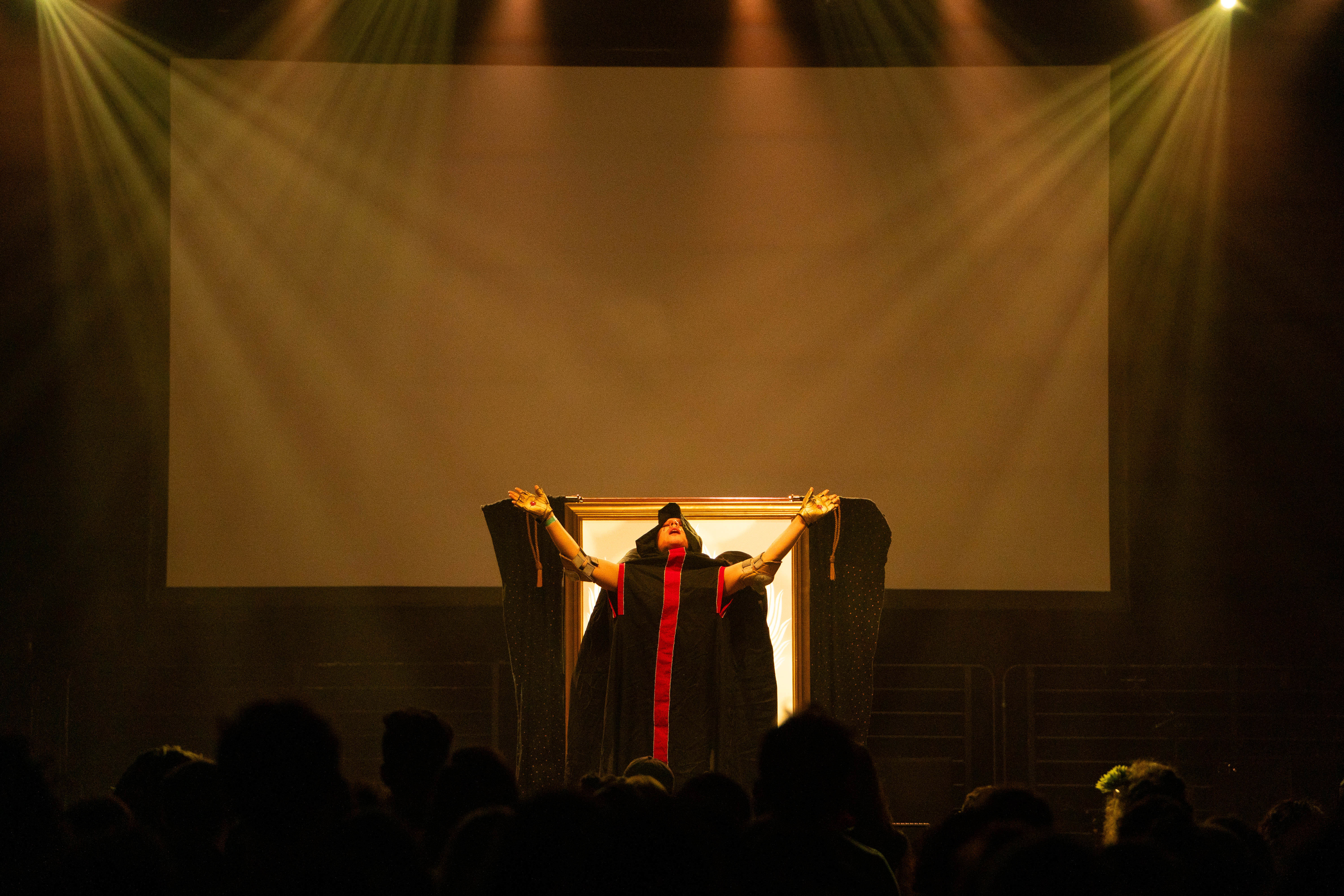 Xtian Mingle stands on stage in a red and black cloak, flinging both hands up.