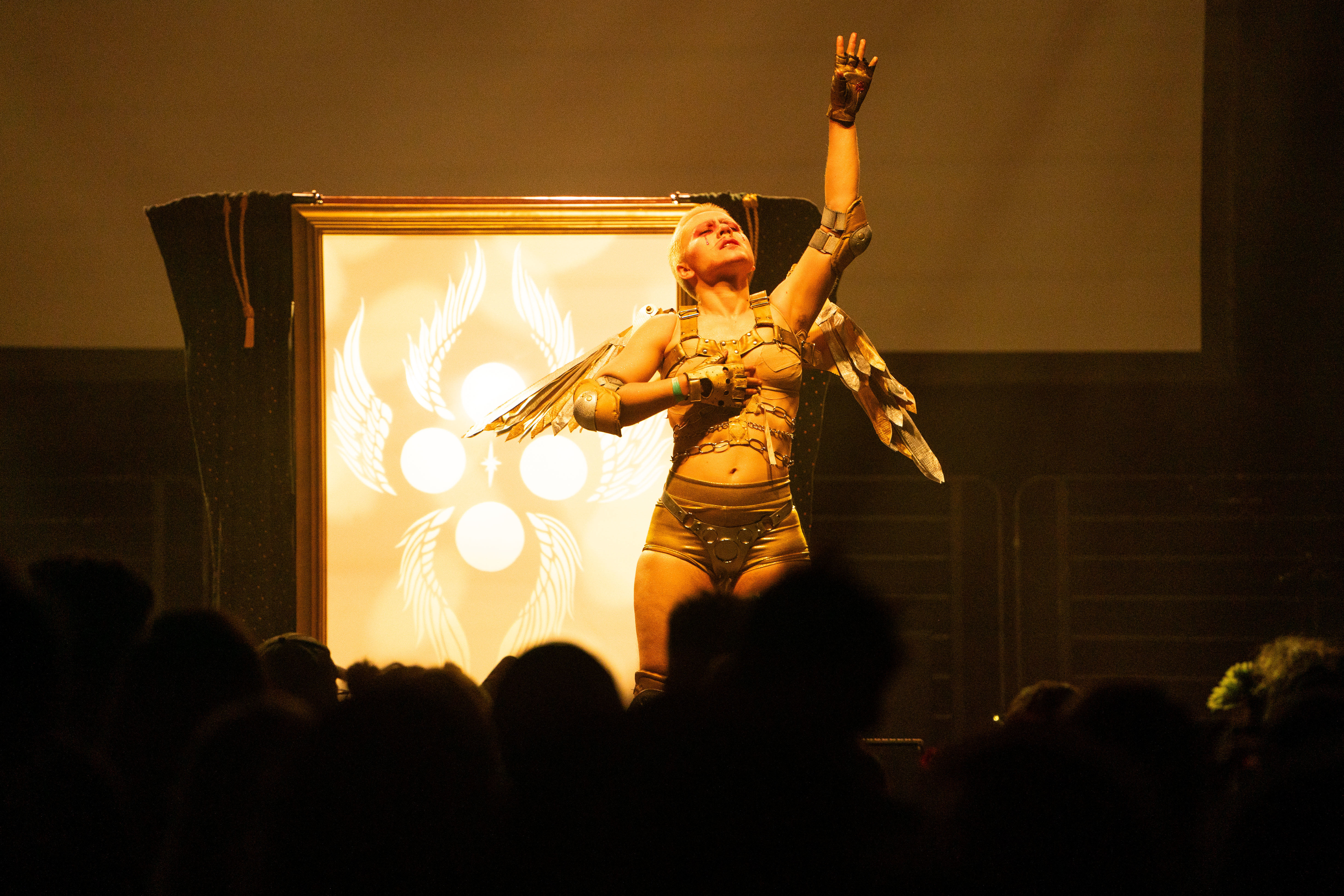 Xtian Mingle onstage in a gold and silver costume with angel wings, reaching upward with one hand.