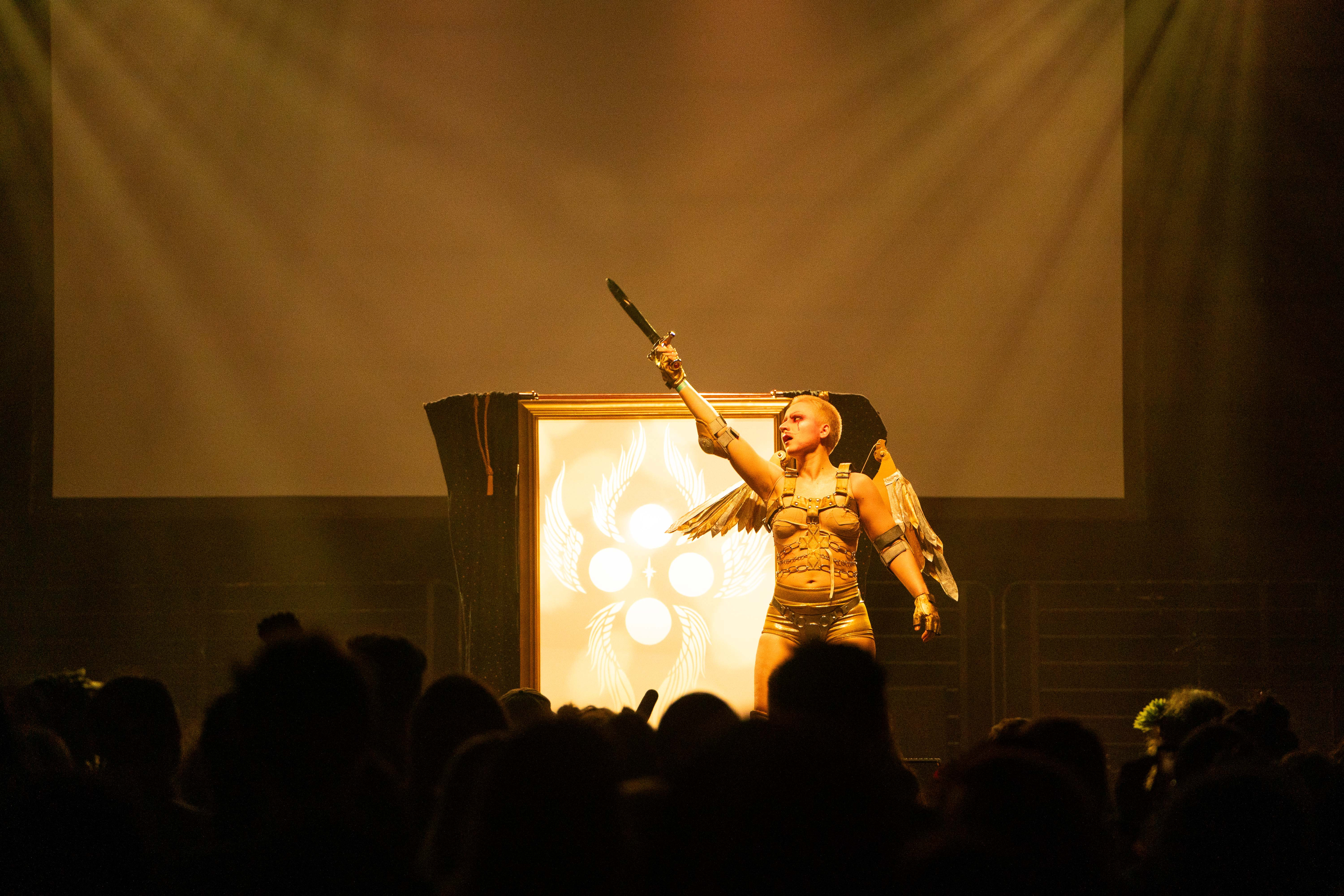 Xtian Mingle onstage, in a gold and silver costume with angel wings, gesturing with a sword.