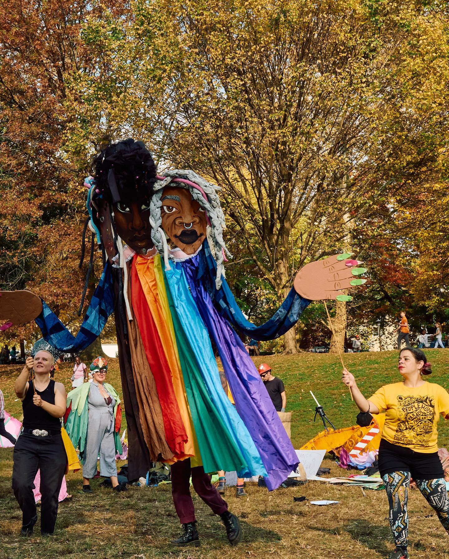 A puppet depicting a 3-faced being in a rainbow dress being carried by 3 people.